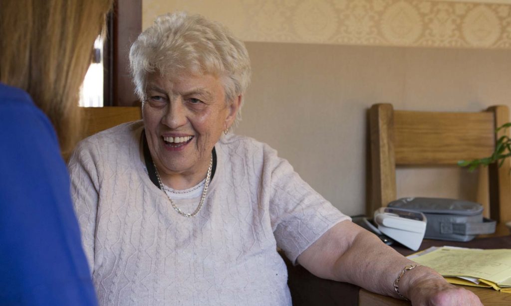 A district nurse with her back to camera talks to an elderly woman in her kitchen