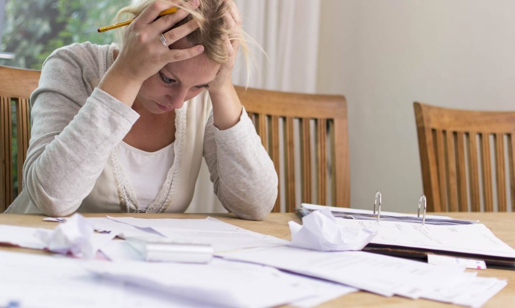 A blond woman with head in hands looks at a table full of bills and invoices