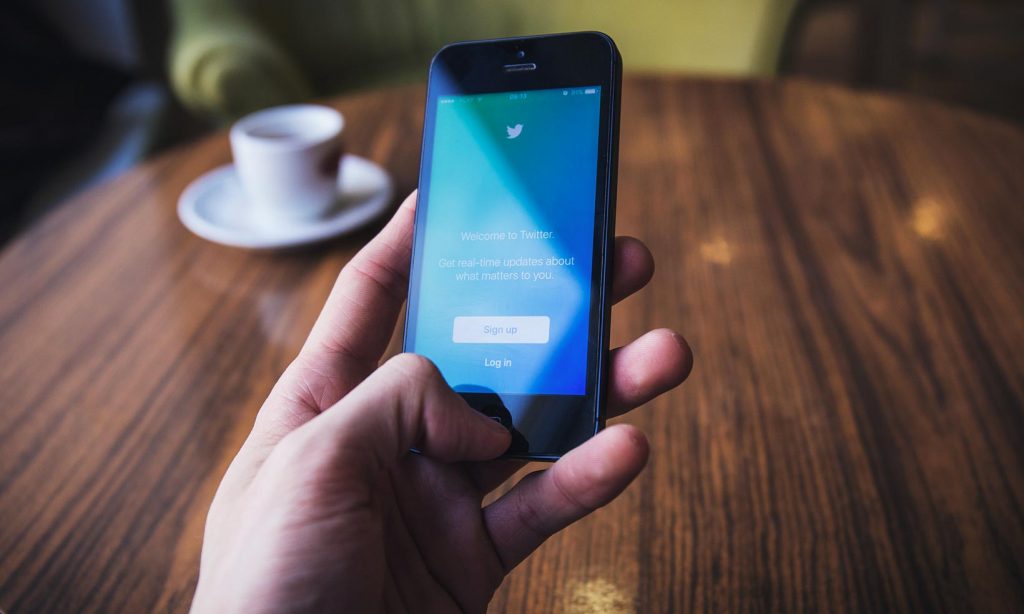 A mans hand holding a mobile phone showing a blue screen with twitter logo
