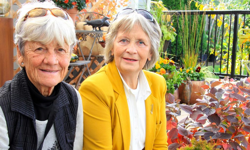 Two retired ladies dressed in bright colours at a garden centre, smiling to camera